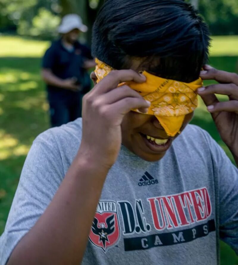 A young person wearing a grey D.C. United Camps t-shirt is blindfolded with a yellow bandana. They are outside in a grassy area, possibly a park, with another person visible in the background wearing a white hat. The blindfolded person is smiling and appears to be adjusting the bandana. The focus is on the individual with the bandana, suggesting they are participating in an outdoor activity or game.